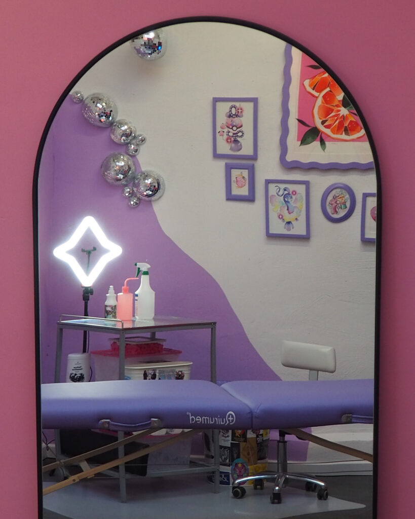Tattoo-Arbeitsplatz vor einer weiß-lila Wellenwand mit glänzenden Discokugeln. Auf dem kleinen Metalltisch stehen Reinigungsmittel, eine Seifenflasche und Tattoo-Zubehör, beleuchtet von einer auffälligen sternförmigen Ringlampe. Darunter befinden sich Stickerboxen mit Statements wie ‚Hot Girl Summer‘ und ‚No Kitsch No Stitch‘ – der ganze Bereich wirkt sauber, kreativ und verspielt.