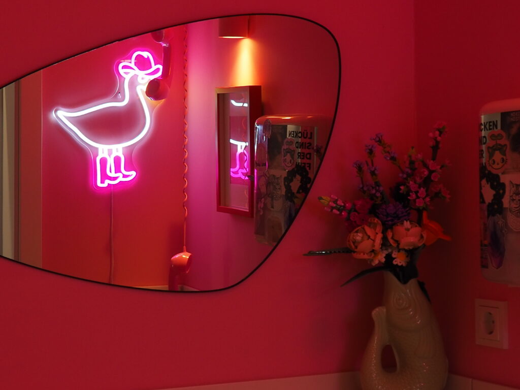 Blick in ein pinkes Badezimmer mit verspieltem Retro-Flair. Im asymmetrischen Spiegel reflektiert sich ein leuchtendes Neonlicht in Form einer Ente mit Cowboyhut und Stiefeln. Darunter hängt ein Vintage-Telefonhörer. Auf der Kommode steht eine Vase in Fischform mit einem bunten Kunstblumenstrauß. Die Wände sind mit Stickern und kleinen Details verziert.