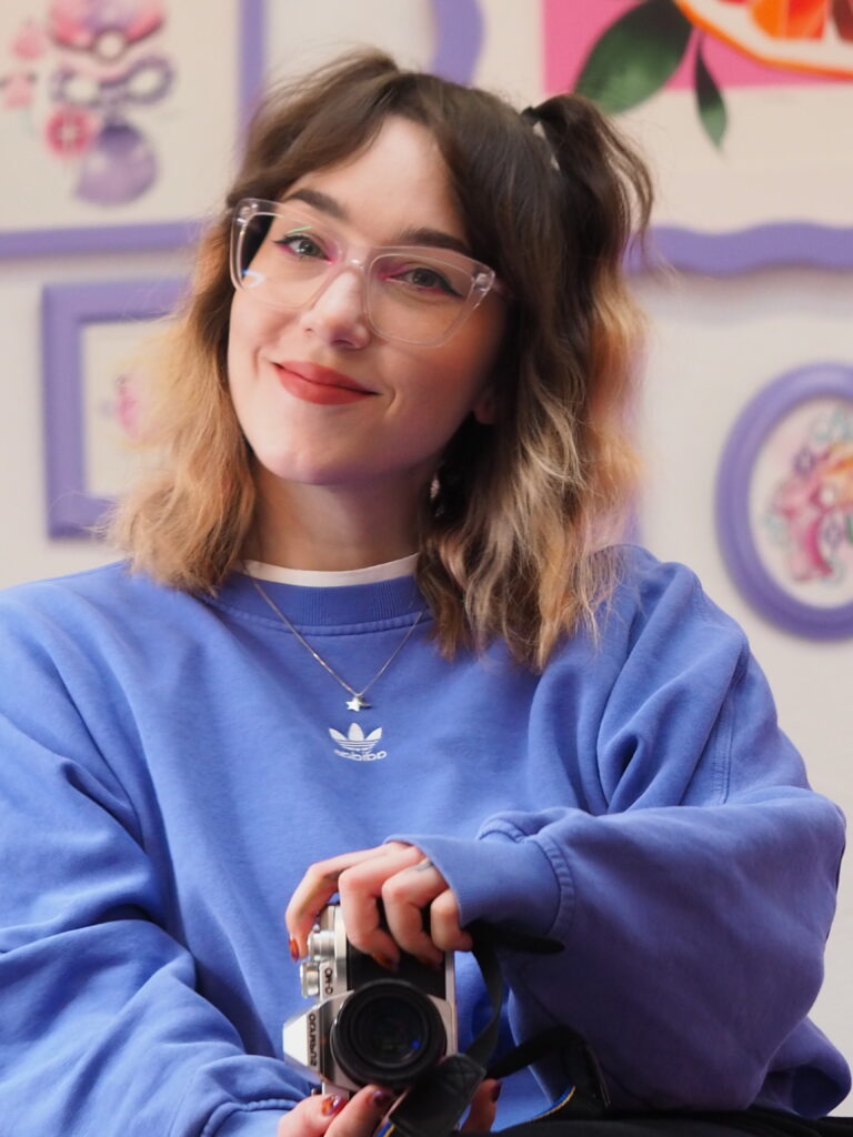 Frau mit Brille und blauer Adidas-Sweatshirt, lächelnd mit Kamera in der Hand, vor pastellfarbenen Illustrationen mit lila Rahmen – Selbstporträt in einem kreativen Atelier