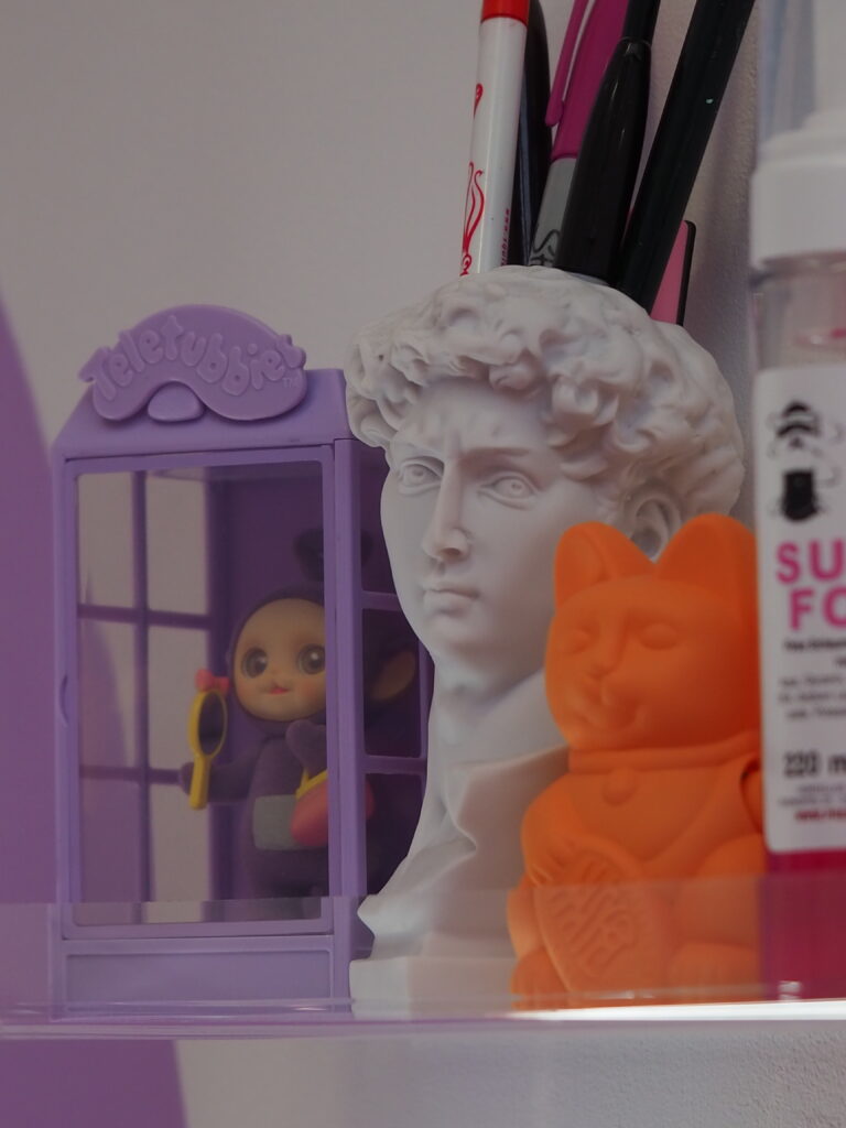 Bunte Kombination aus Dekofiguren und Zeichenmaterialien auf einem Regal: ein lila Teletubbies-Spielhäuschen mit Tinky Winky-Figur, eine weiße David-Büste als Stifthalter, ein orangefarbener Winkekatze-Dekoartikel sowie bunte Marker und Stifte. Im Hintergrund: ein Teil einer Flasche mit Tattoo-Zubehör. Kreativ, kitschig und charmant inszeniert.