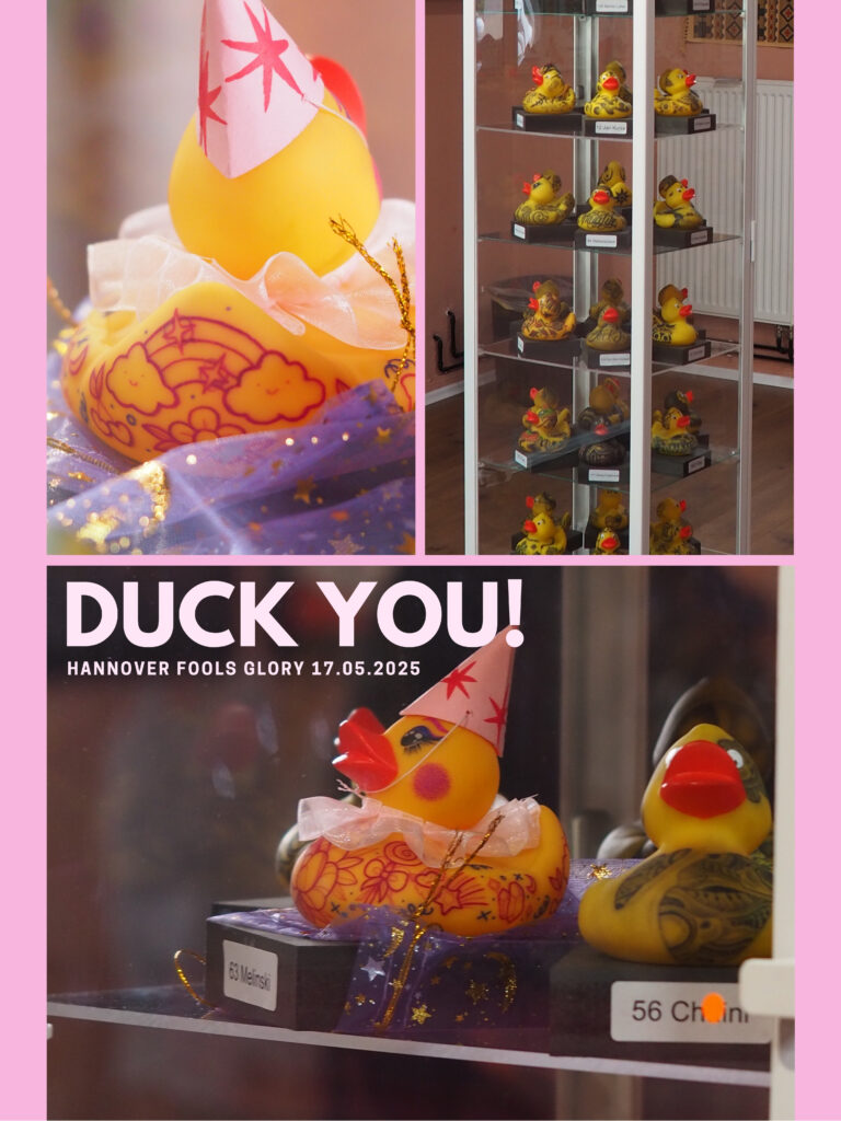 Ausstellungsansicht mit künstlerisch gestalteten Badeenten in einer Vitrine, einige mit Partyhut und floralen Tattoos, ergänzt durch den Schriftzug ‚Duck You! – Hannover Fools Glory 17.05.2025