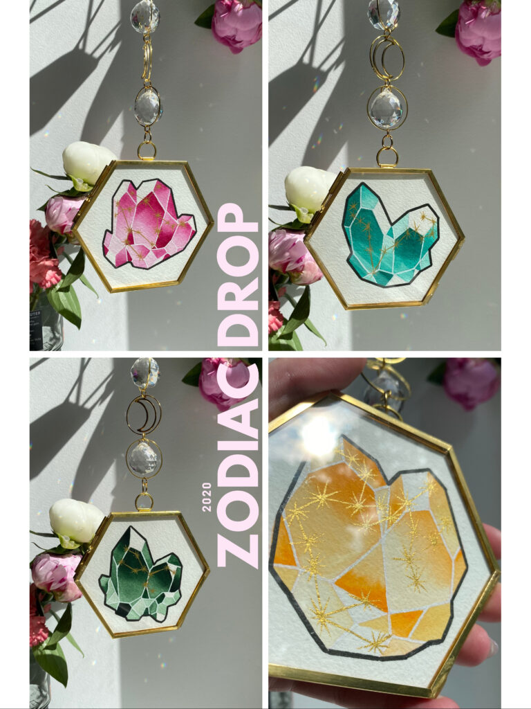 Handgefertigte Sonnenfänger mit gemalten Kristallmotiven in Rosa, Grün und Gelb, eingefasst in goldene sechseckige Rahmen, aufgehängt im Sonnenlicht mit Blumen im Hintergrund.