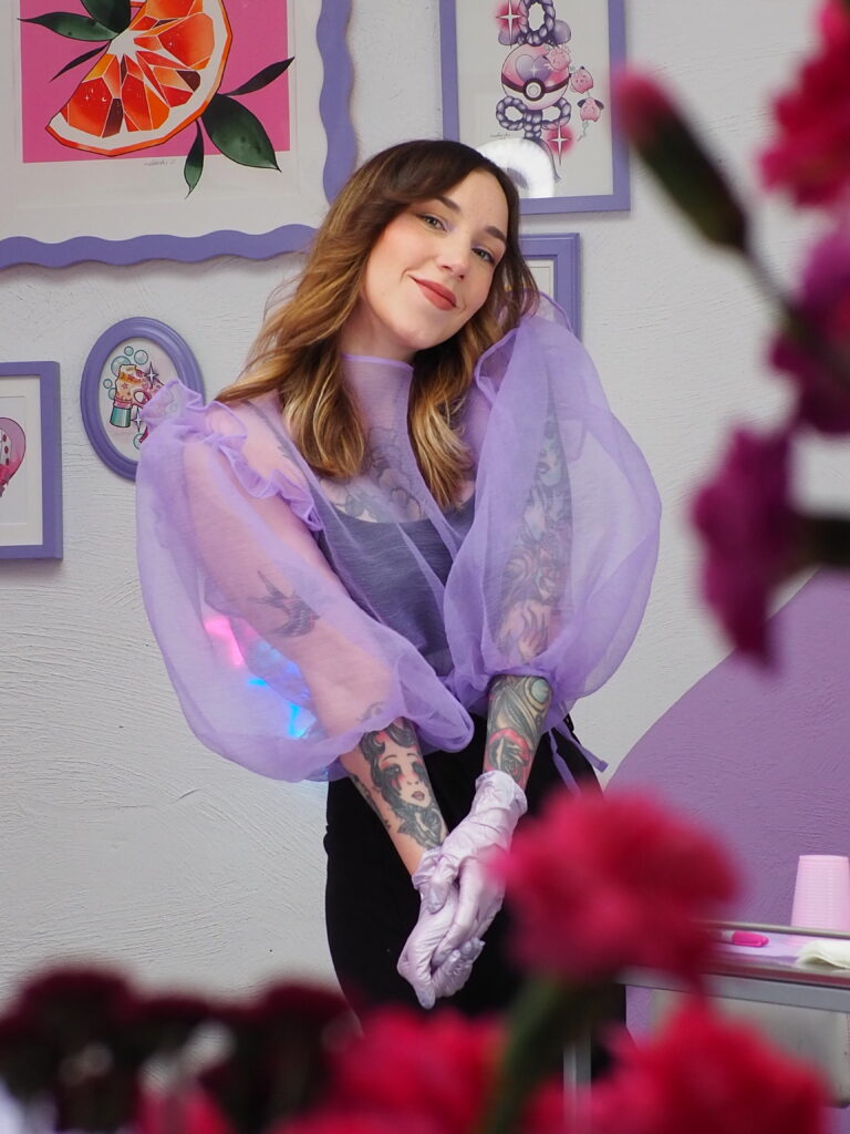 Porträt einer Künstlerin in einem lilafarbenen Atelier, die lächelnd in die Kamera blickt. Sie trägt eine transparente, puffärmelige Bluse in Flieder, Einweghandschuhe und steht vor gerahmten Illustrationen, im Vordergrund sind unscharfe pinke Blumen zu sehen